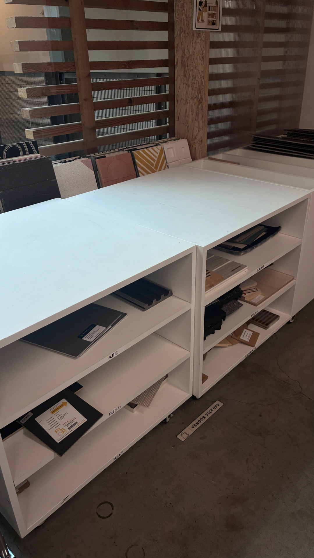 Materials library shelving unit with alphabetically labeled shelves holding sample boards, beside a Vendor Pickups floor marker